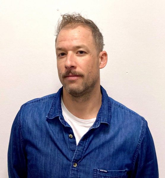 Portrait of a man with short messy hair and a mustache, wearing a blue denim shirt over a white T-shirt, standing against a plain white background.