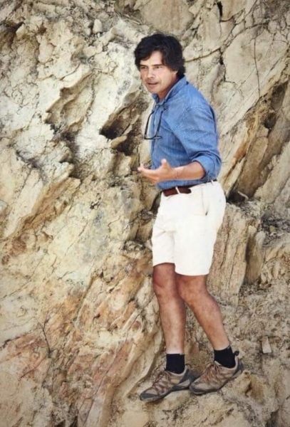 A man in a blue long-sleeve shirt, white shorts, black socks, and hiking shoes standing against a rocky, textured cliff face, gesturing with his right hand and wearing glasses hanging on his shirt collar.