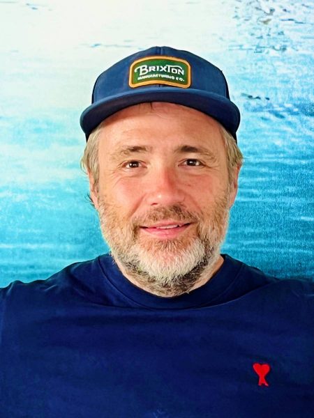 Middle-aged man with a light beard wearing a navy blue Brixton cap and a navy blue shirt with a small red heart emblem, standing against a blurred blue background.
