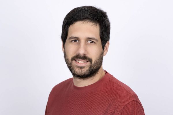 Portrait of a man with dark hair and beard wearing a red sweater against a light background.