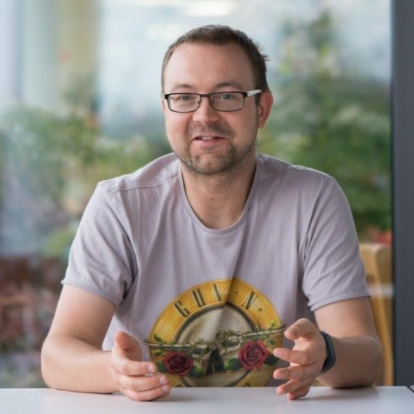 Man with glasses wearing a light purple Guns N' Roses t-shirt, seated at a table with a blurred outdoor background.
