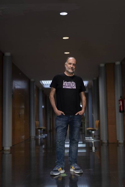A middle-aged man with a beard standing in a hallway with reflective black flooring, wearing a black t-shirt with white text that reads 'PERIFERIAS RESURRECTION,' blue jeans, and gray sneakers with yellow details, hands in pockets, under round ceiling lights.