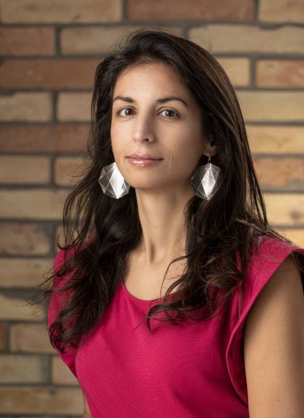 Portrait of a woman with long dark hair wearing large geometric silver earrings and a bright pink sleeveless top, standing against a blurred brick wall background.
