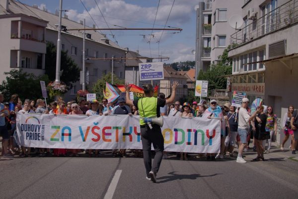 A crowd of people participating in a pride march in an urban street, holding a large banner that says 'DUHOVY PRIDE BRATISLAVA ZA VŠETKY RODINY' with a person in a yellow vest and megaphone standing in front of them raising their hands, surrounded by colorful flags and signs supporting equality and LGBTQ+ rights.
