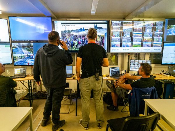 A control room with multiple large screens displaying surveillance footage and event monitoring; three men are present, two standing and one sitting, monitoring the screens closely, one man talking on a phone and another wearing a radio device on his belt.