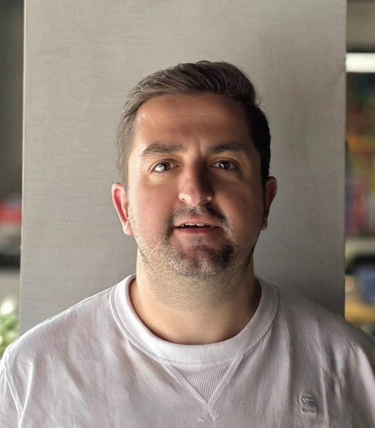 Portrait of a man with short dark hair and light beard wearing a white sweatshirt, standing against a plain gray background indoors.