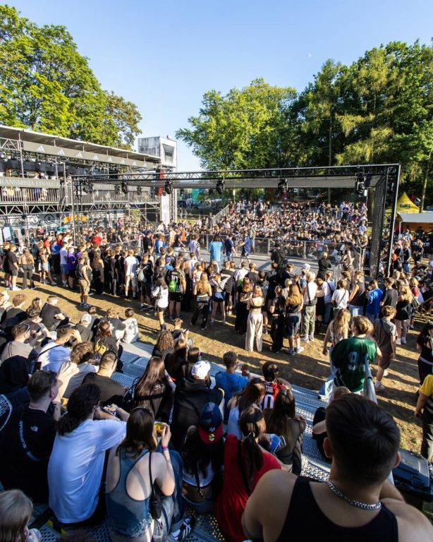 Large crowd of people watching an outdoor event inside a fenced cage in a park with trees and blue sky.