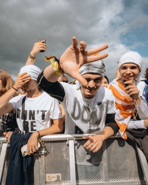 Three young men standing behind a metal barricade at an outdoor event, wearing white head coverings and casual clothes, one making a hand gesture towards the camera, another holding a microphone and wrapped in an orange and white flag, with a cloudy sky in the background.