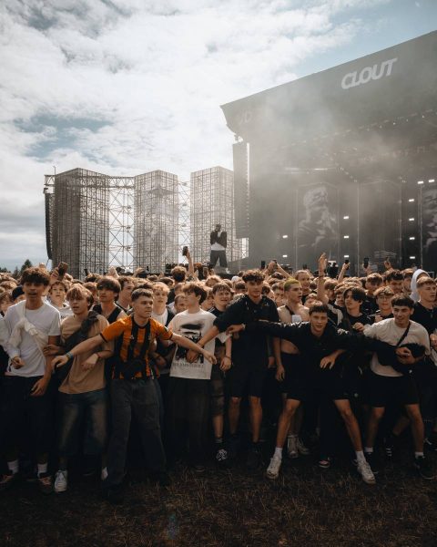 Crowd of young people standing close together at an outdoor music festival with a performer on stage in the background under a cloudy sky.