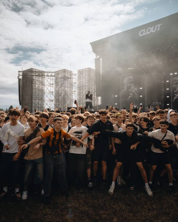 Crowd of young people standing close together at an outdoor music festival with a performer on stage in the background under a cloudy sky.