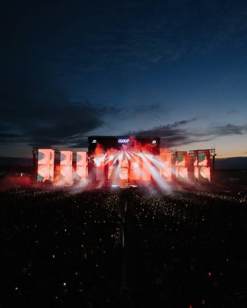Large outdoor concert stage lit with bright red and white lights and smoke effects at dusk, with a massive crowd holding up lights in the foreground.