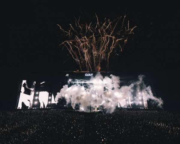 Large outdoor concert stage at night with fireworks exploding overhead, smoke effects on stage, bright lights, and a massive crowd in front