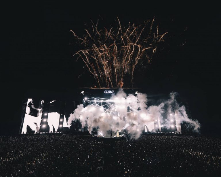 Large outdoor concert stage at night with fireworks exploding overhead, smoke effects on stage, bright lights, and a massive crowd in front