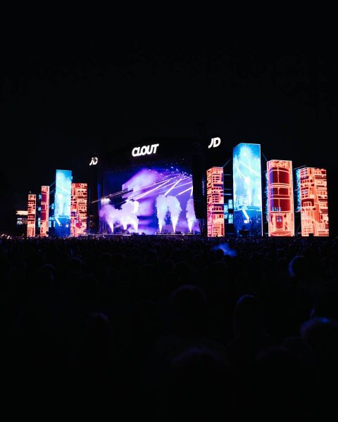 Large outdoor music concert stage at night with bright colorful LED screens in red and blue patterns and white smoke effects, with a crowd of people in front of the stage under dark sky