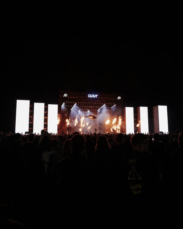Crowd gathered at an outdoor nighttime concert with a large stage displaying the word 'CLOUT', bright vertical screens on either side, stage lights, flames, and a helicopter flying above the stage.