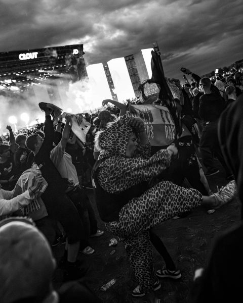 Black and white photo of a lively outdoor concert crowd with people cheering and raising their hands, one person in the foreground wearing a leopard print outfit energetically dancing or jumping, with a large stage in the background under a cloudy sky.
