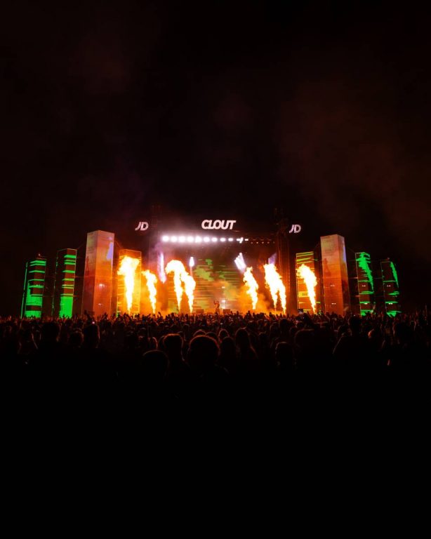 Large crowd at a nighttime music concert with a stage featuring bright flames and illuminated green pillars, with the word 'CLOUT' displayed above the stage.