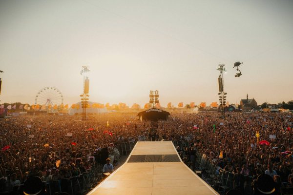 Large crowd gathered at an outdoor music festival at sunset with stage platform extending towards audience and a ferris wheel visible in the background