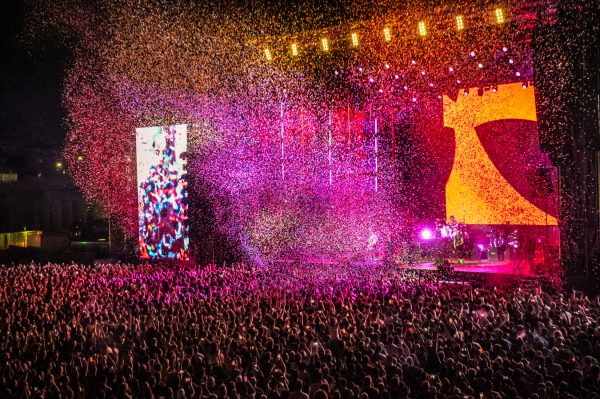 Large crowd at a night concert with colorful confetti falling over the audience and bright stage lights illuminating the performers and screens.