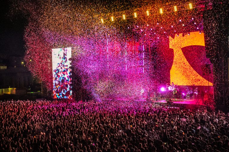 Large crowd at a night concert with colorful confetti falling over the audience and bright stage lights illuminating the performers and screens.