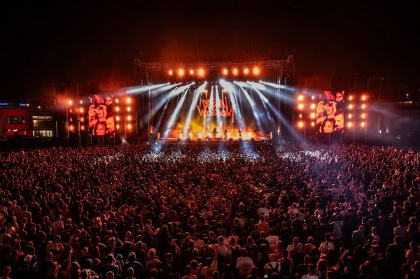 Large crowd gathered at an outdoor concert at night with a brightly lit stage and the words 'Open Wisdom' displayed on a screen behind performers.