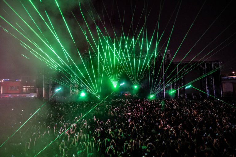 Large crowd at a nighttime outdoor concert with green laser lights projecting from the stage into the sky, many attendees holding up phones to record the event.