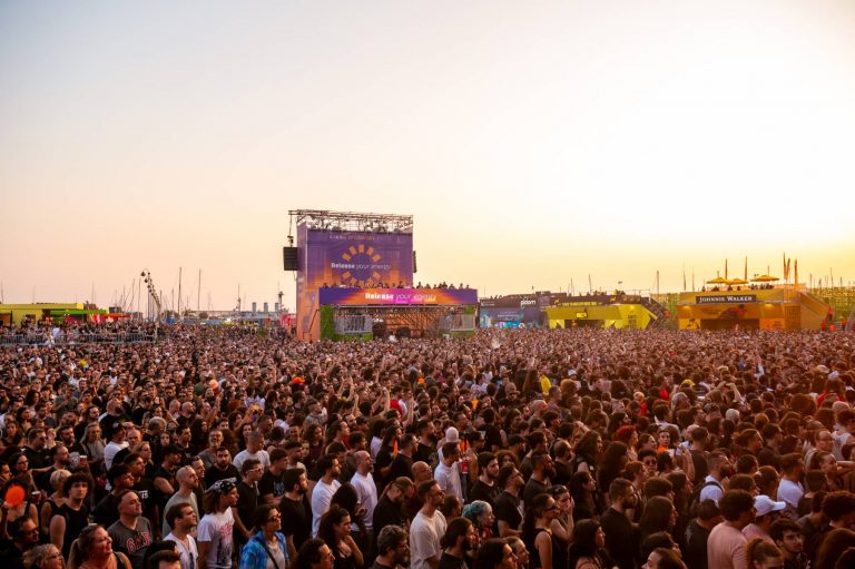 A large crowd of people gathered outdoors at a music festival or concert during sunset, with banners and stages in the background displaying logos and phrases like 'Release your energy' and 'Johnnie Walker'.