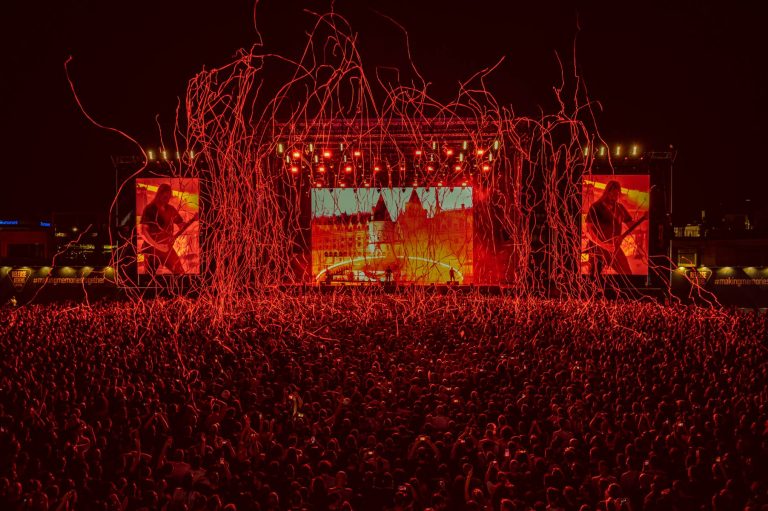 Large crowd at a nighttime outdoor concert with a brightly lit stage and red streamers flying through the air, surrounded by large screens showing the performer playing a guitar.