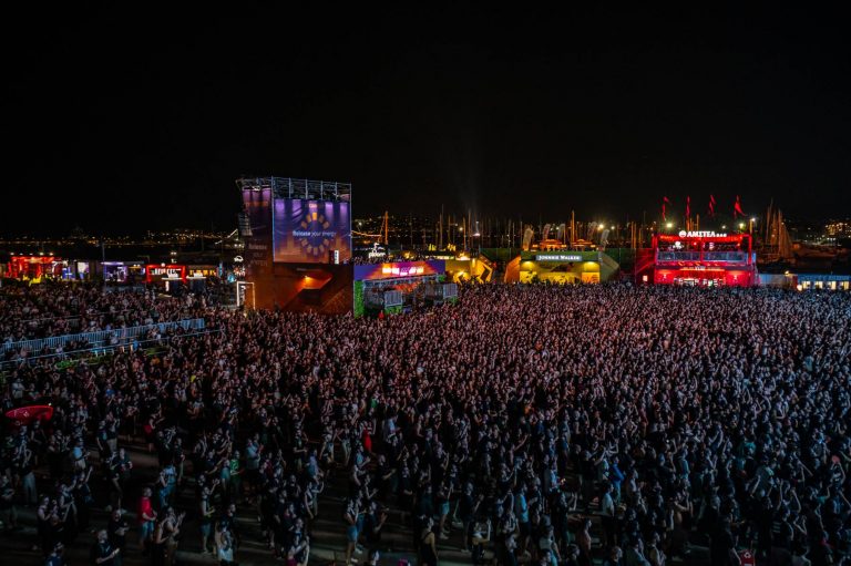 Large crowd of people gathered outdoors at night for a concert or festival with illuminated sponsor booths and structures in the background under a dark sky.