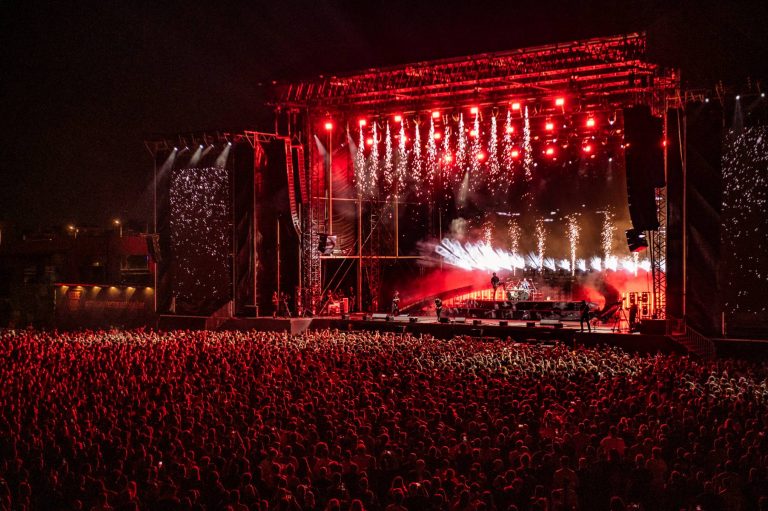 Large crowd illuminated by red lights at a nighttime outdoor concert with a stage featuring red lighting, pyrotechnics, and performers playing music.