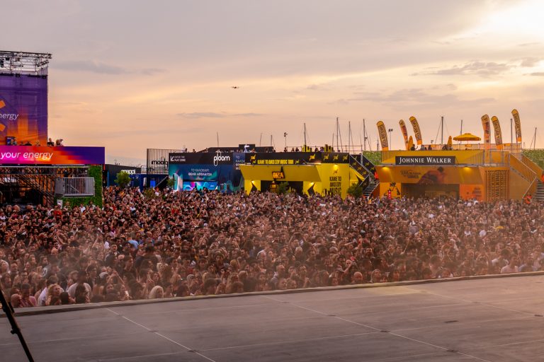 A large crowd of people gathered at an outdoor event during sunset with promotional booths and banners for Johnnie Walker and other brands in the background, and a stage in the foreground.