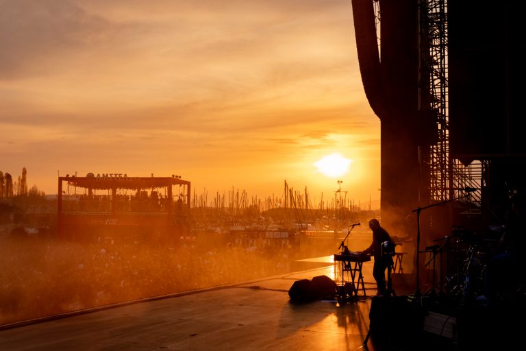Silhouetted musician performing on an outdoor stage during a golden sunset with a crowd and marina filled with sailboat masts in the background.