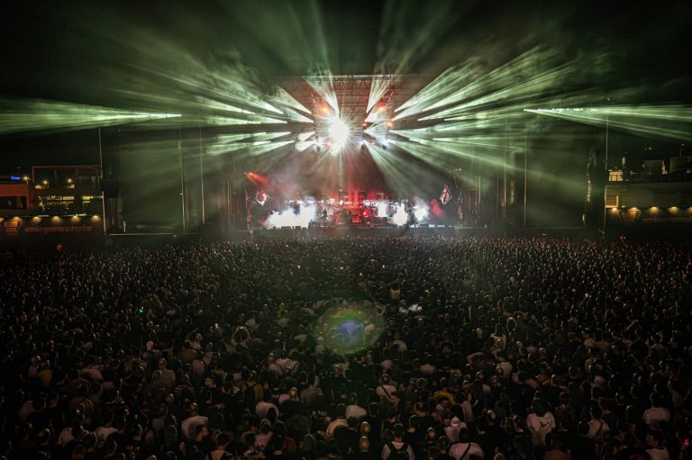 A large crowd at a night concert with bright stage lights and fog effects illuminating the band performing on stage.