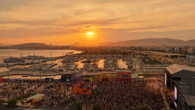 A large outdoor concert with a dense crowd in front of a stage near a marina filled with boats at sunset, with mountains and a city skyline in the background.
