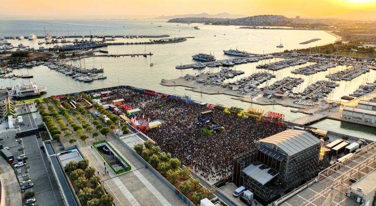 A large outdoor music festival with a densely packed crowd in front of a large stage near a marina filled with boats at sunset.