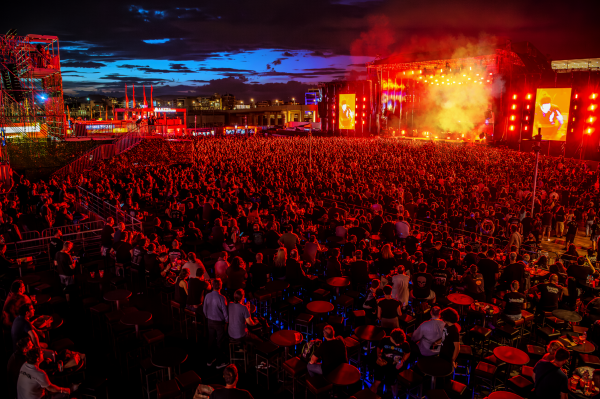 Large outdoor concert at night with a densely packed crowd facing a stage illuminated in red lights and smoke, against a dark blue sky at dusk.