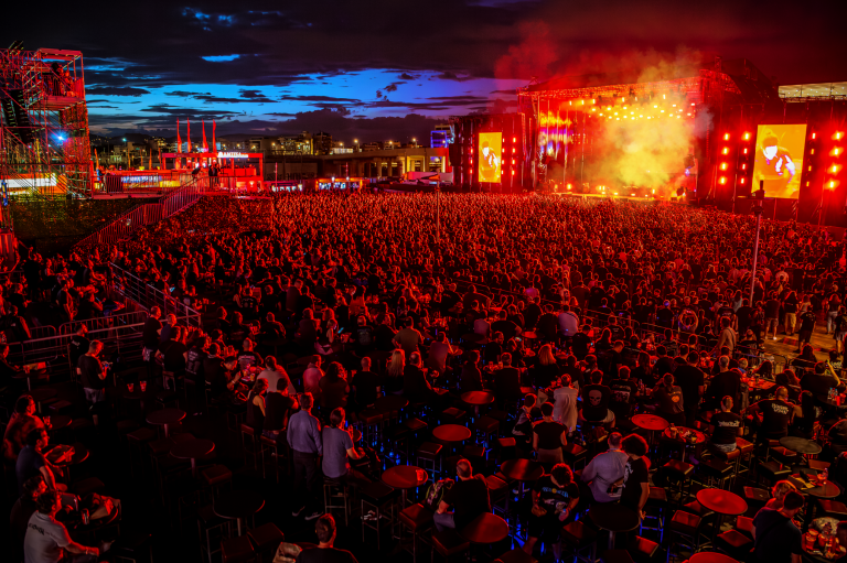 Large outdoor concert at night with a densely packed crowd facing a stage illuminated in red lights and smoke, against a dark blue sky at dusk.