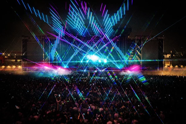 Large outdoor concert at night with a crowd facing a stage illuminated by vibrant blue and purple laser lights creating geometric patterns.