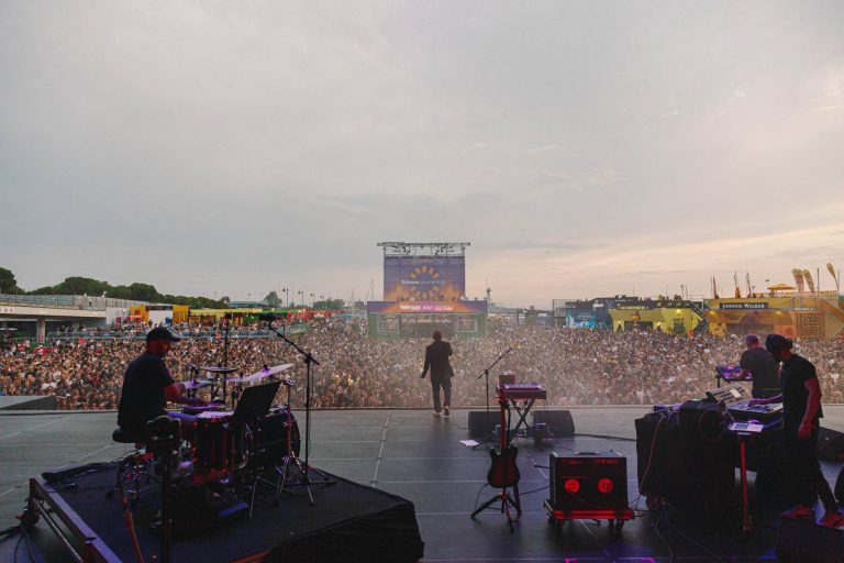 View from a concert stage showing a drummer, two keyboard/synth players, and a singer facing a large crowd of people under a cloudy evening sky with stage equipment visible in the foreground.