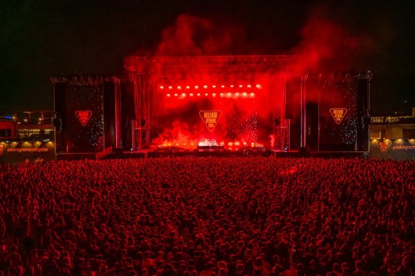 Large crowd illuminated by red lights facing an outdoor concert stage with a 