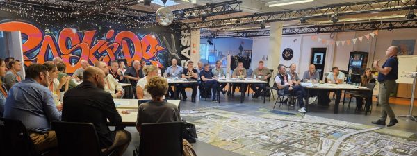 Group of people seated around tables in a large room with graffiti art on the wall and a detailed city map spread out on the floor, listening to a standing speaker giving a presentation.