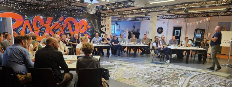 Group of people seated around tables in a large room with graffiti art on the wall and a detailed city map spread out on the floor, listening to a standing speaker giving a presentation.