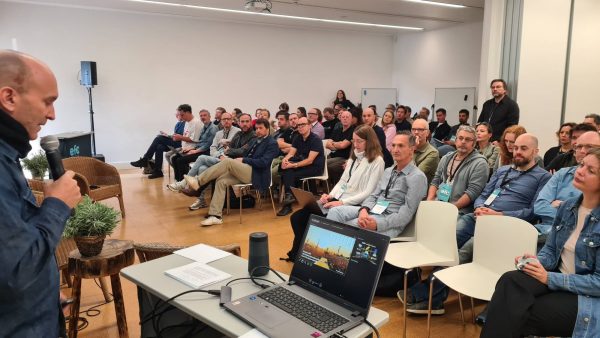 Man speaking into microphone in front of an audience seated in a conference room with a laptop showing a presentation on the table.