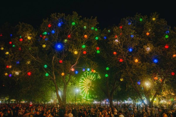 A large crowd of people gathered at a night festival with trees decorated with colorful lanterns and a brightly lit Ferris wheel in the background.