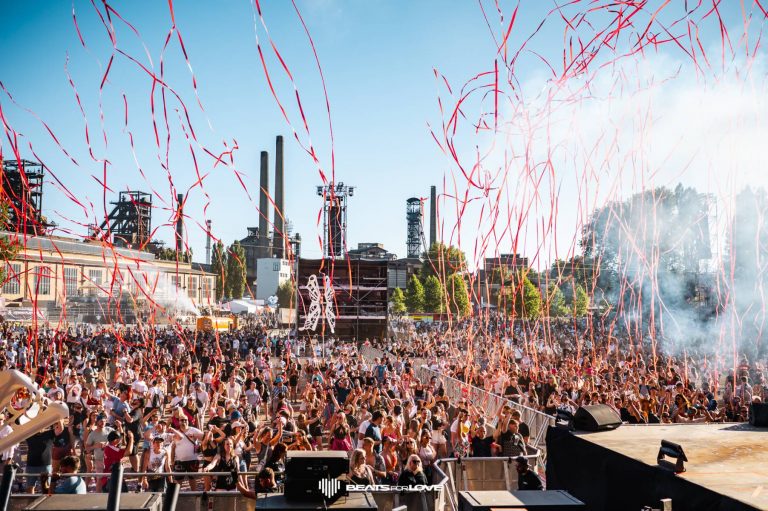 Large outdoor music festival crowd celebrating with red streamers flying above and stage smoke on a clear day.