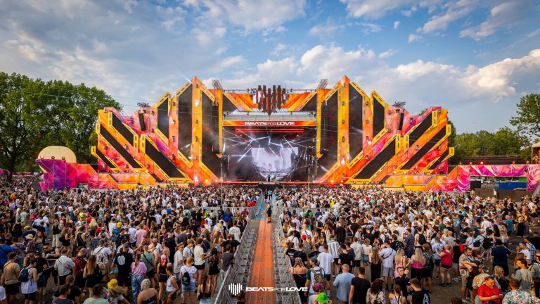 Large outdoor music festival crowd facing a bright orange and black geometric stage with 