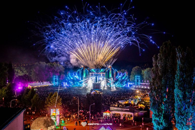 Nighttime music festival with a large crowd, a brightly lit stage, colorful lights, and fireworks exploding overhead