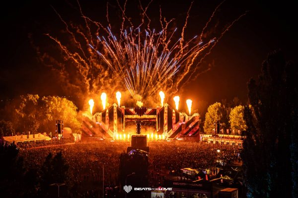 Nighttime electronic music festival with a large crowd, illuminated stage, fire effects, and fireworks overhead.