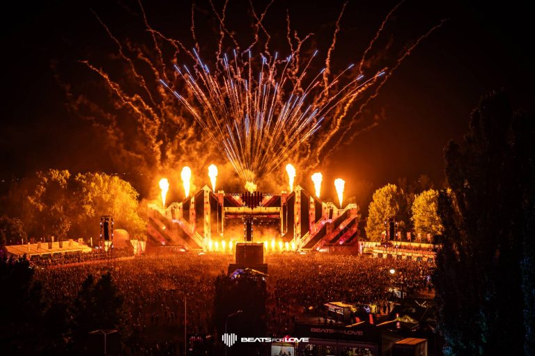 Nighttime electronic music festival with a large crowd, illuminated stage, fire effects, and fireworks overhead.