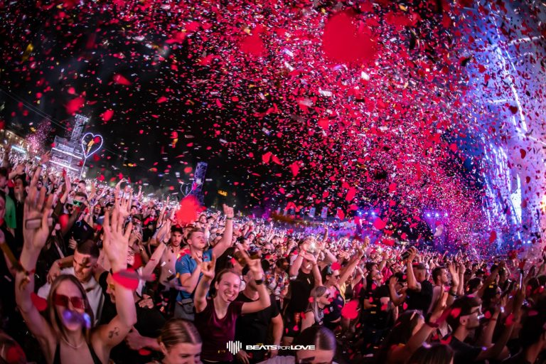 Large energetic crowd celebrating at an outdoor night music festival with red confetti filling the air.